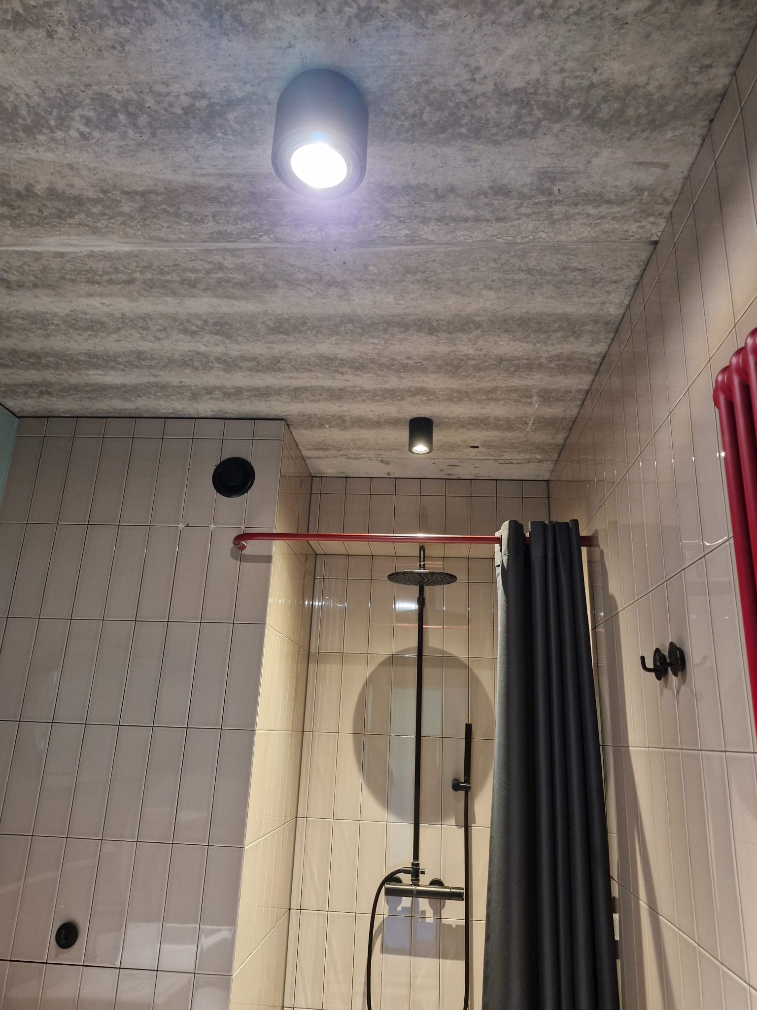 Nowoczesna łazienka z beżowymi płytkami, betonowym sufitem i czarną armaturą. Widoczny prysznic z deszczownicą i szara zasłona. Minimalistyczny design.