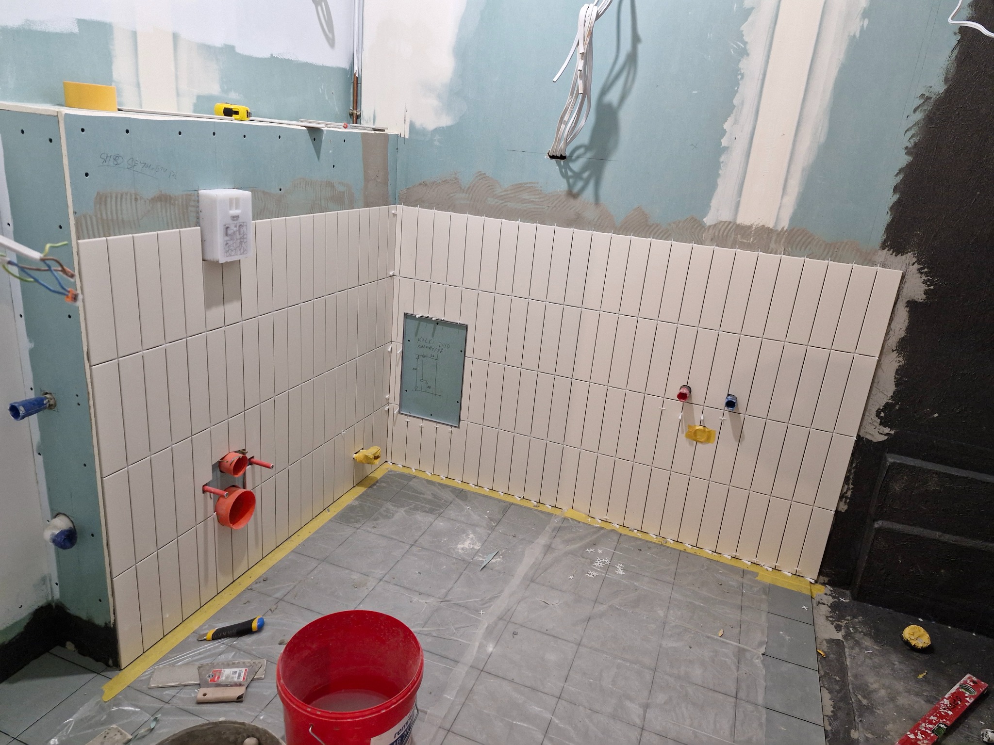 Białe, pionowo ułożone płytki na ścianie w łazience w trakcie remontu. Widoczne instalacje wodne, narzędzia glazurnicze i zabezpieczona folią podłoga z szarych płytek.
