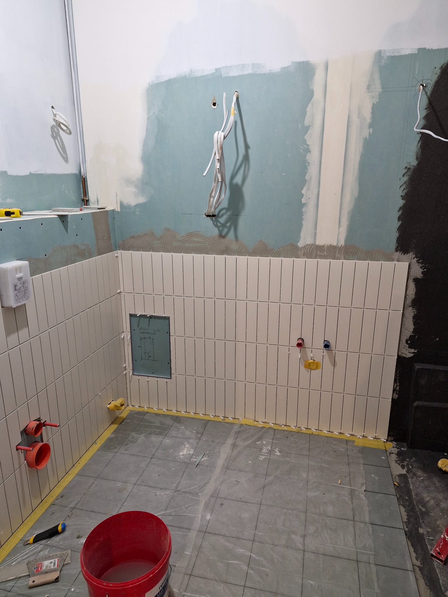 Łazienka w trakcie remontu: ułożone pionowe płytki w odcieniach bieli na ścianie, widoczne instalacje, przygotowane podłoże pod posadzkę. Częściowo wykończone wnętrze.