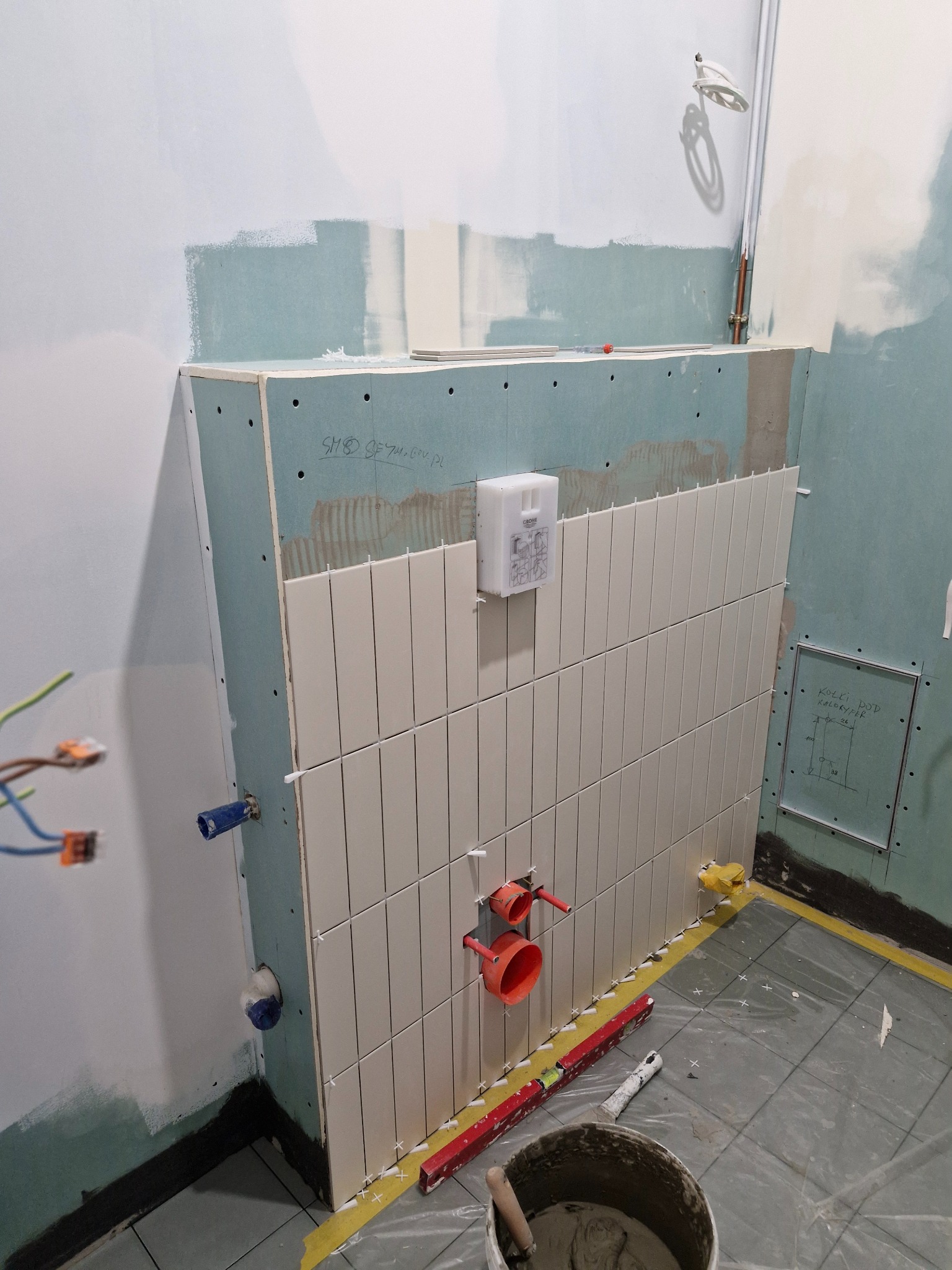 Białe, wąskie płytki układane pionowo na ścianie w Gdyni. Widoczne elementy instalacji, poziomica i wiadro z zaprawą. Prace glazurnicze w trakcie realizacji.