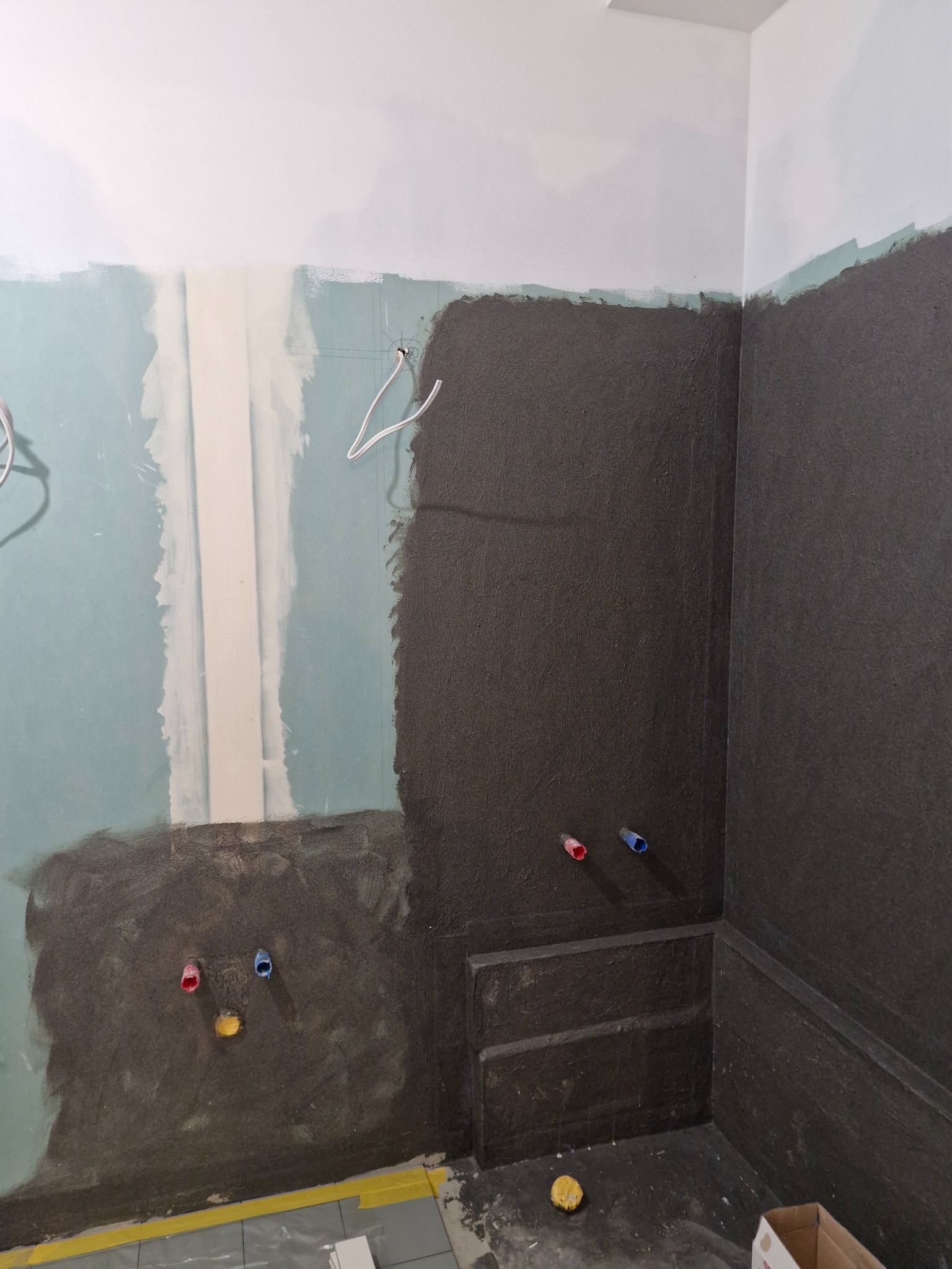 Hydroizolacja w łazience w trakcie prac: ściany pokryte ciemną masą, widoczne rury instalacyjne, fragmenty płyt gipsowych. Przygotowanie do dalszych prac wykończeniowych.