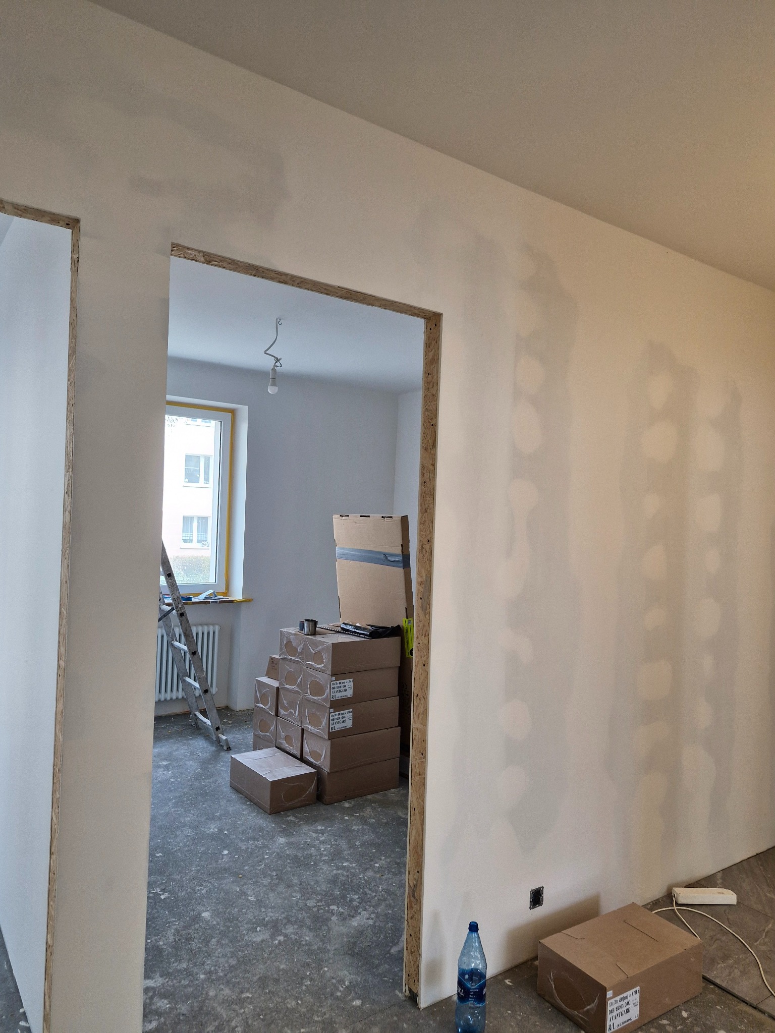 Ściana po szpachlowaniu, widoczne ślady po łatach. W tle okno z żółtą ramą, drabina i kartony. W pomieszczeniu widać butelkę wody i gniazdko elektryczne.