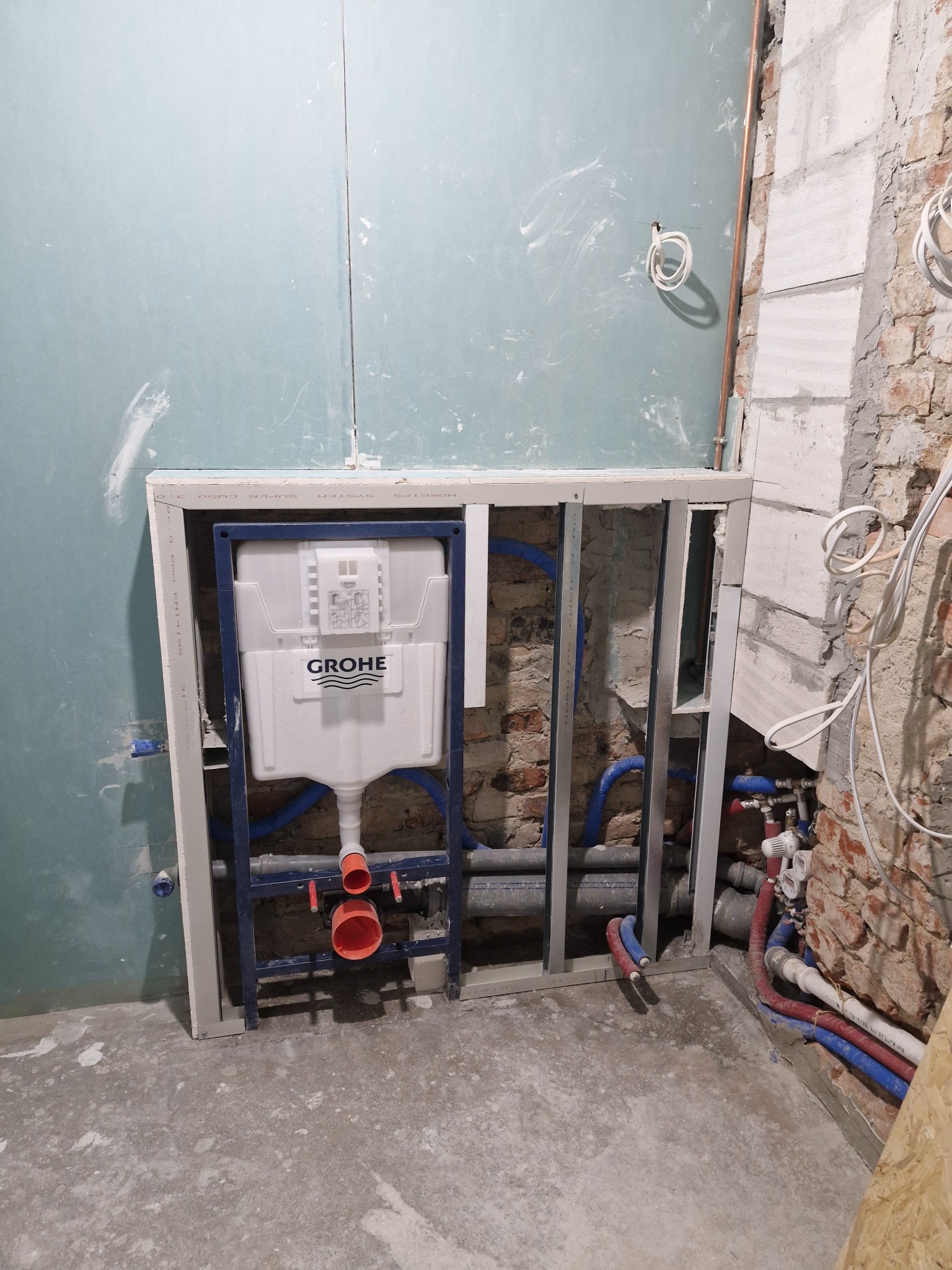 Montaż stelaża podtynkowego Grohe z instalacją wodno-kanalizacyjną na tle surowych ścian z cegły i płyty gipsowej. Widoczne niebieskie i czerwone rury.