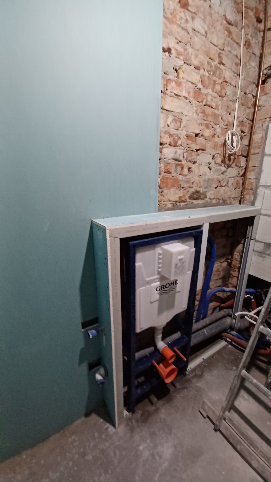 Instalacja podtynkowa WC Grohe w obudowie z płyt gipsowo-kartonowych na tle surowej cegły i fragmentu ściany z płyt GK. Widoczne rury instalacyjne.