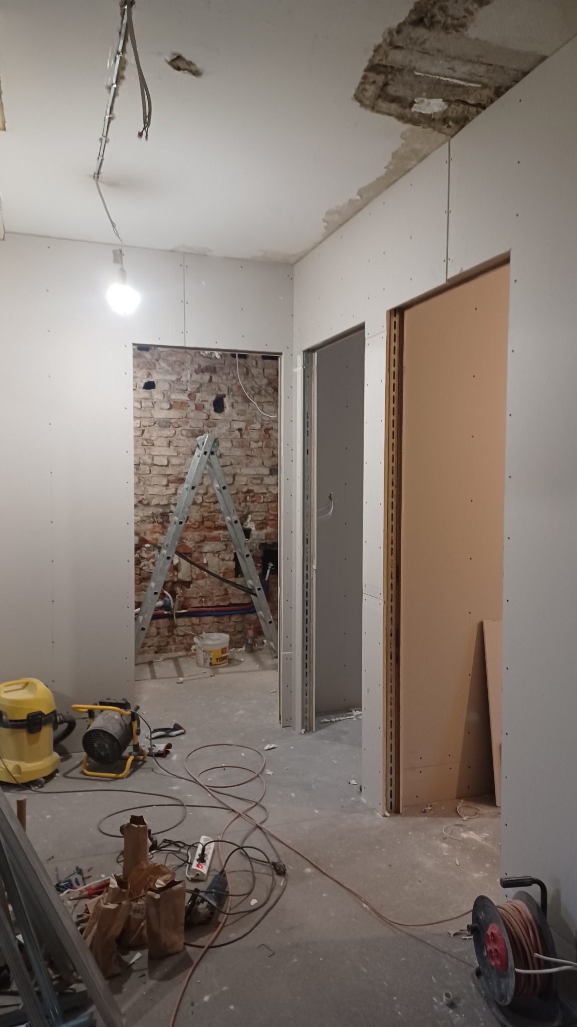 Remont wnętrza: ściany z płyt gipsowo-kartonowych, widoczna cegła w przejściu, drabina, narzędzia budowlane. Prace w toku, surowy stan wykończenia.