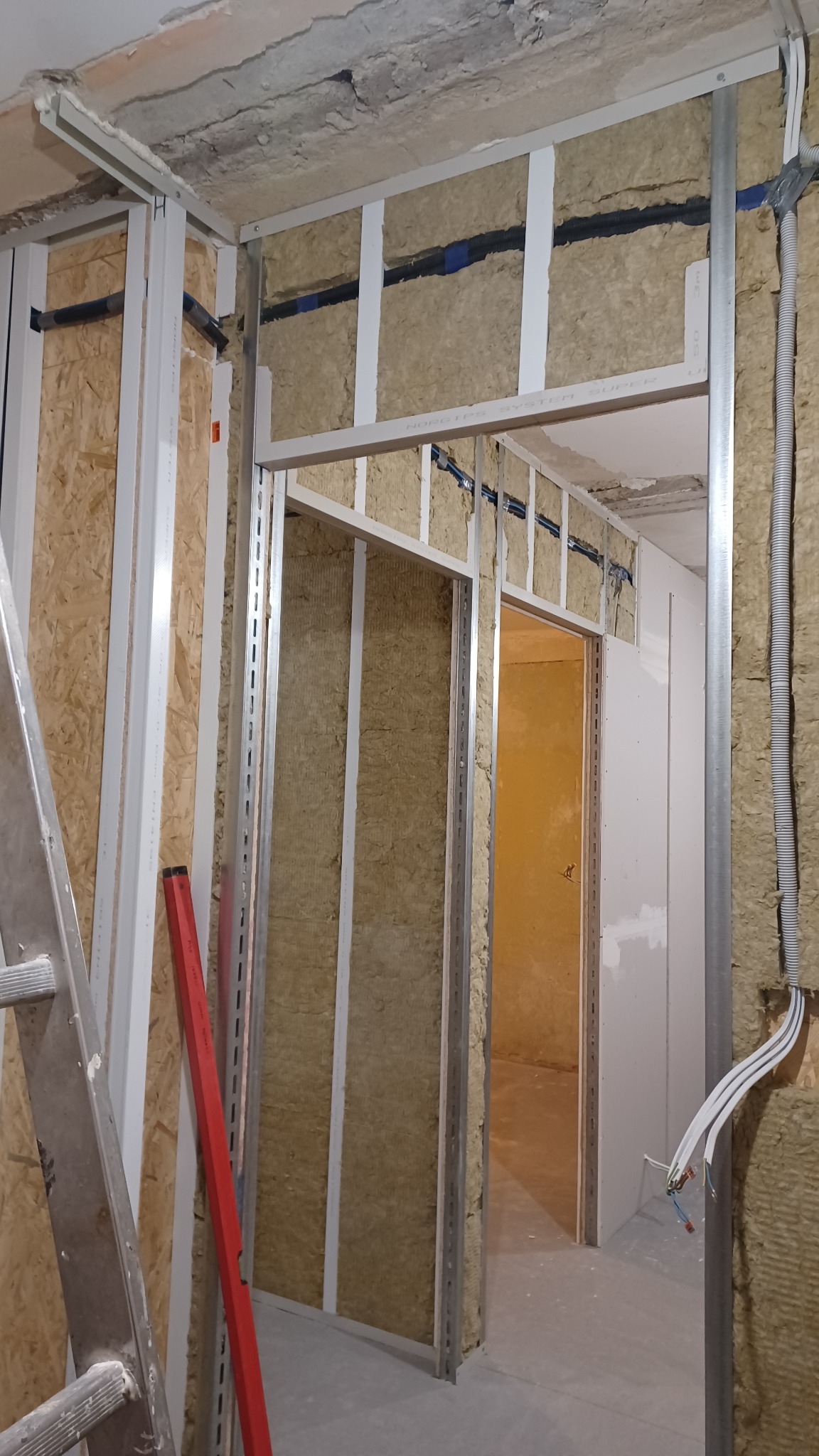 Montaż ścian działowych z izolacją z wełny mineralnej i metalowym stelażem. Widoczne kable elektryczne. Czerwona poziomica oparta o ścianę.