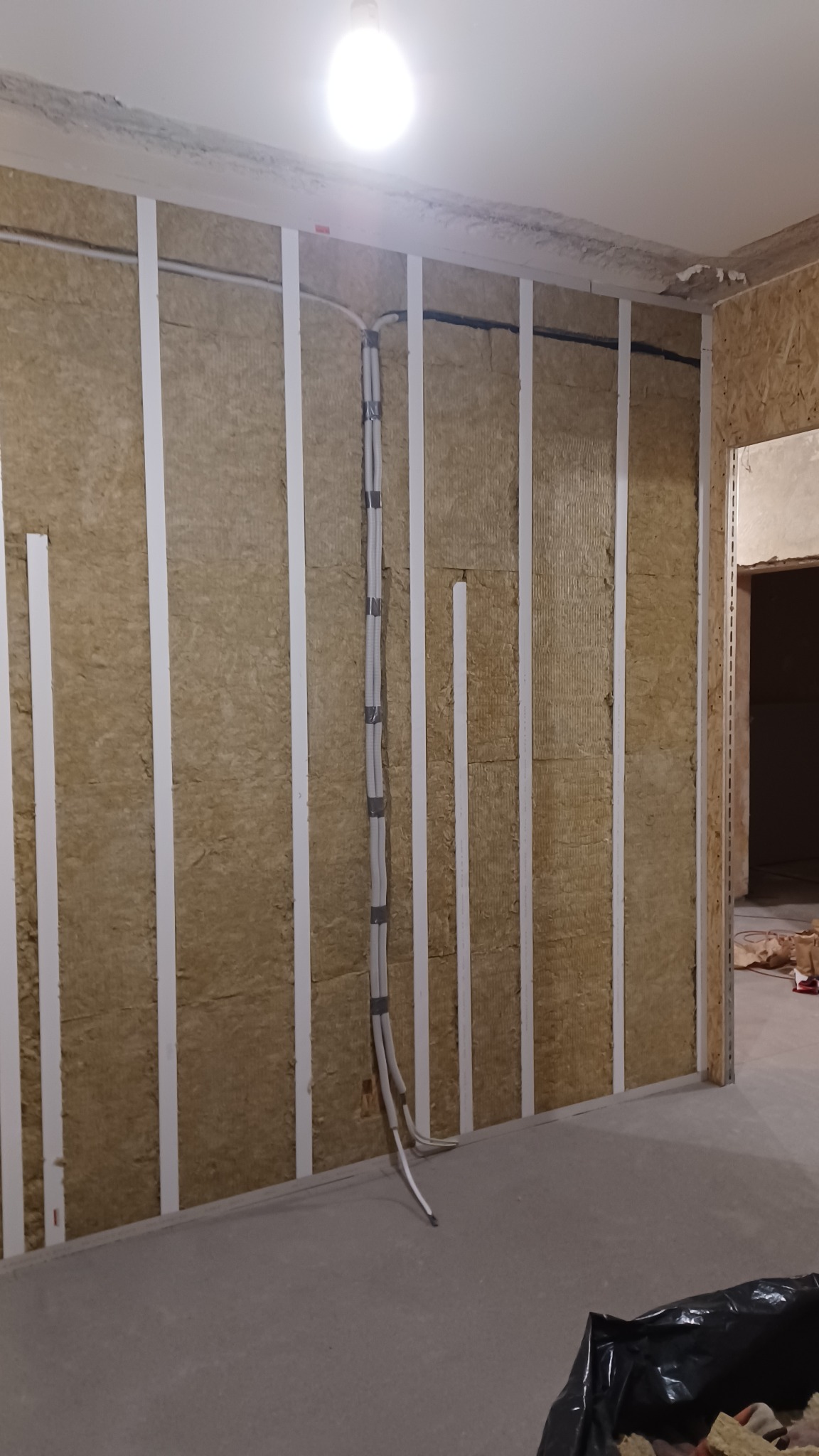 Ocieplenie ściany wełną mineralną z widocznymi przewodami elektrycznymi, zamocowanymi do ściany za pomocą taśmy. Widoczne białe listwy montażowe.