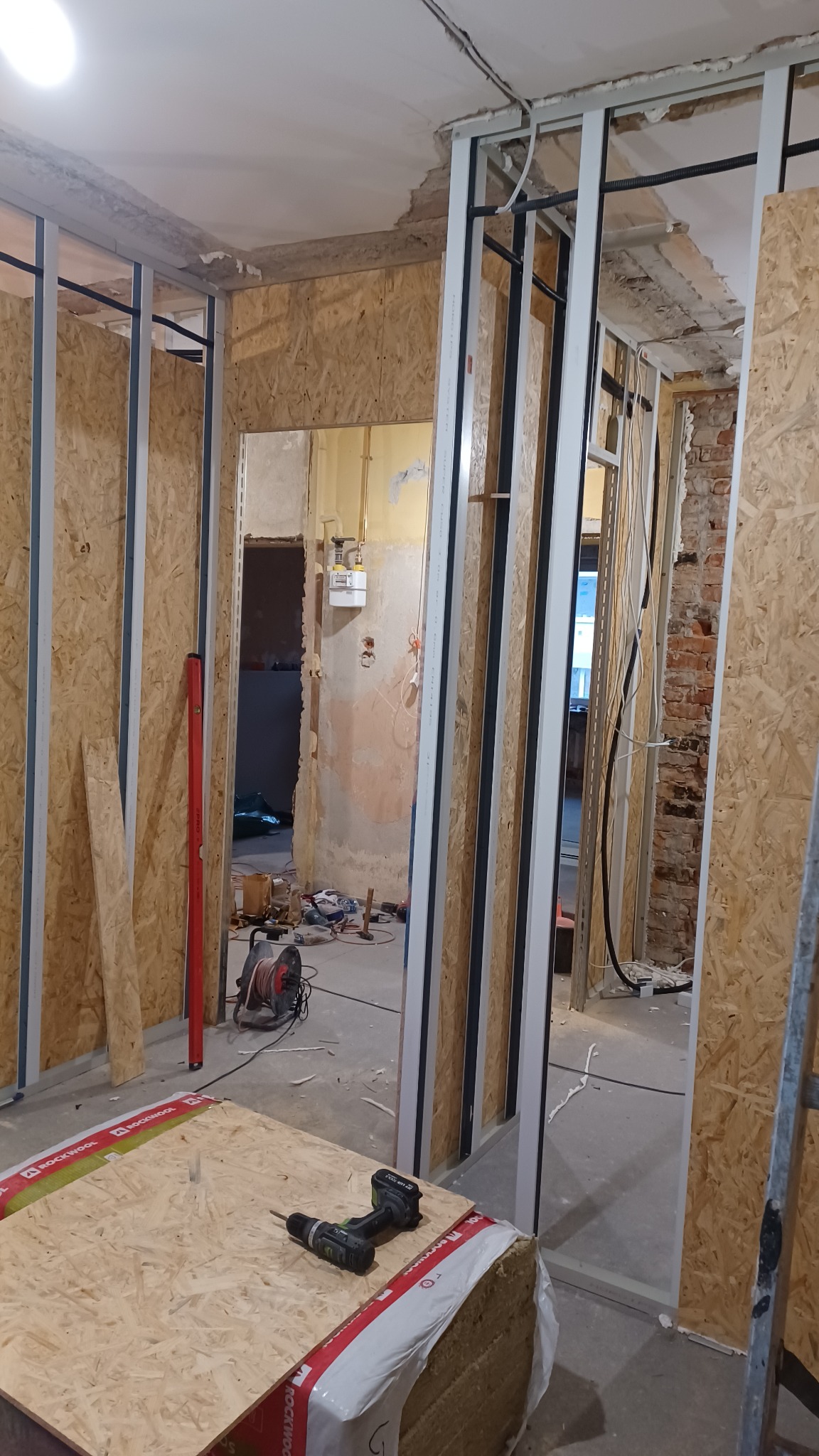 Remont mieszkania w trakcie: widoczne stelaże ścian z płyt OSB, narzędzia (wiertarka) i materiały izolacyjne. Przewody elektryczne, surowy stan ścian w tle.