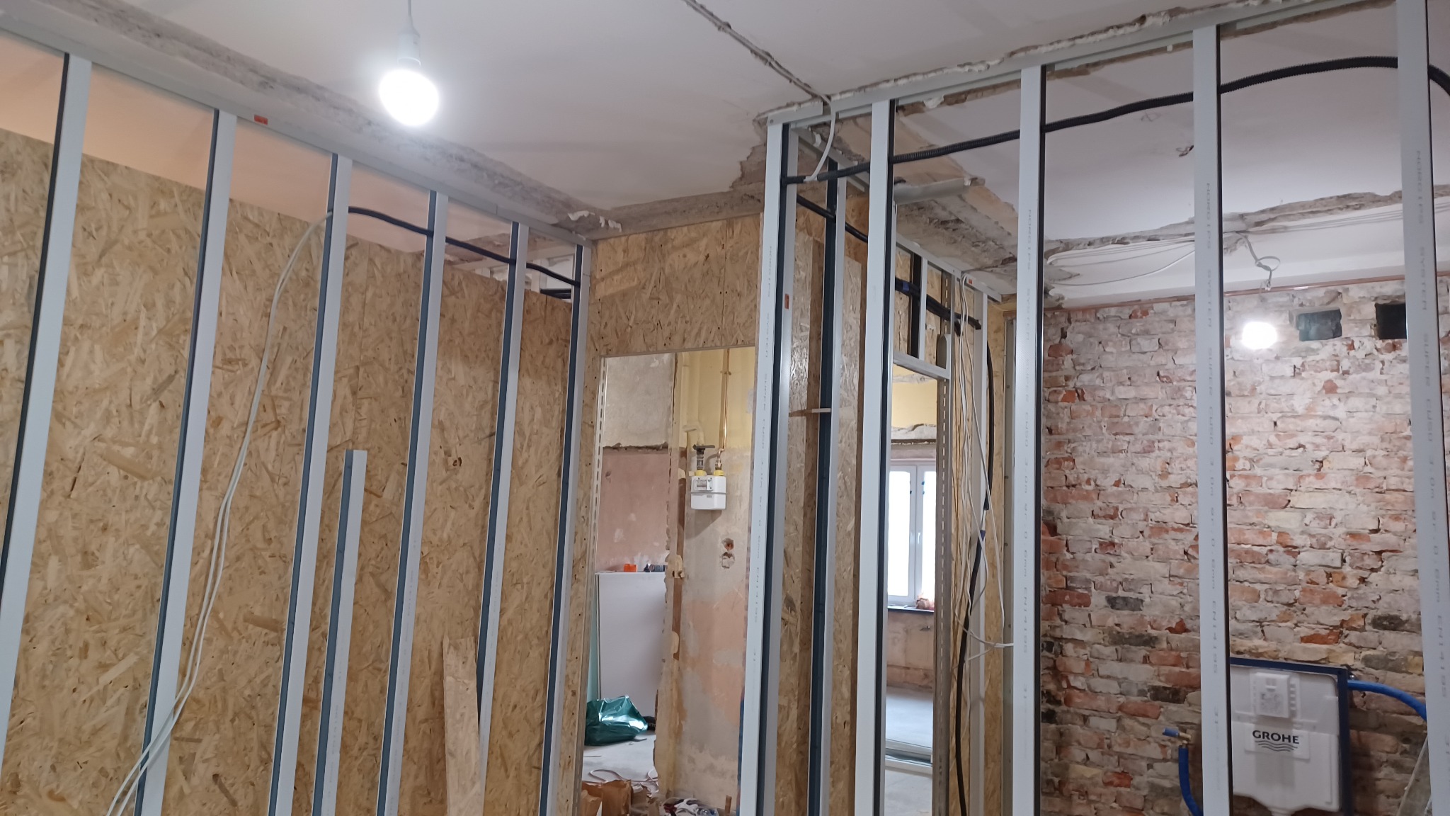 Wnętrze w trakcie remontu: widoczne metalowe profile konstrukcyjne na ścianach z płyt OSB i cegły, instalacje elektryczne, surowy sufit. Etap prac wykończeniowych.