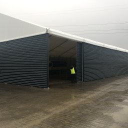 Przedsiębiorstwo Usług Produkcji i Handlu ECOPOL Ireneusz Zioła - Hala z płyt warstwowych w szary odcieniu, z otwartymi wrotami, przez które widać zaparkowany samochód dostawczy. Dwóch pracowników w kamizelkach odblaskowych.