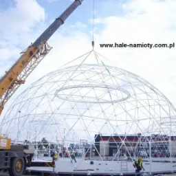 Żuraw budowlany podnosi element konstrukcyjny kopuły geodezyjnej na placu budowy, widoczni pracownicy w kamizelkach odblaskowych, w tle budynek z czerwonym dachem i adres strony internetowej...