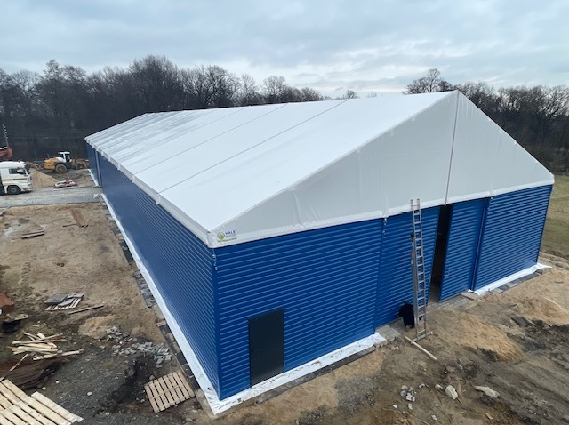 Niebieska hala z białym dachem, widok z góry. Na zewnątrz drabina oparta o ścianę, w tle drzewa i sprzęt budowlany. Hala z blachy falistej.