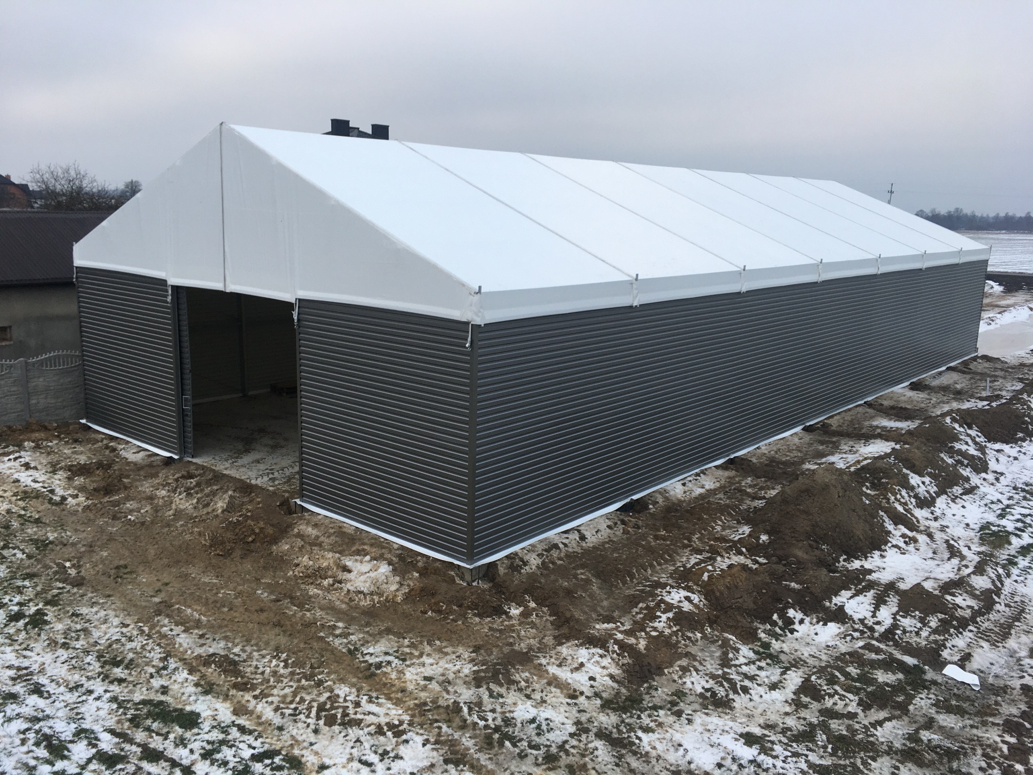 Gotowa hala z szarej blachy falistej i białym dachem z plandeki, widoczna z zewnątrz na tle zimowego krajobrazu. Otwarta brama wjazdowa.