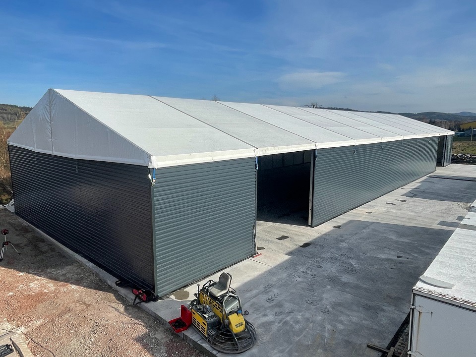 Gotowa hala z szarej blachy falistej i białym dachem, widok z góry. Na pierwszym planie zacieraczka do betonu. Otwarta brama wjazdowa. Słoneczny dzień.