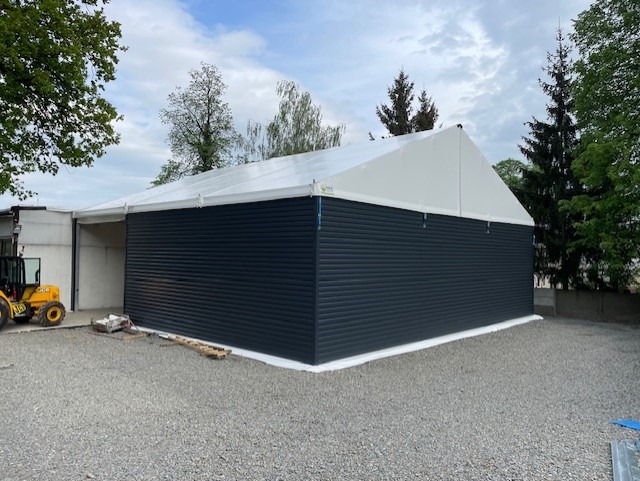 Hala z ciemnej blachy trapezowej i białym dachem z plandeki, widoczny fragment koparki, teren utwardzony żwirem, drzewa w tle.