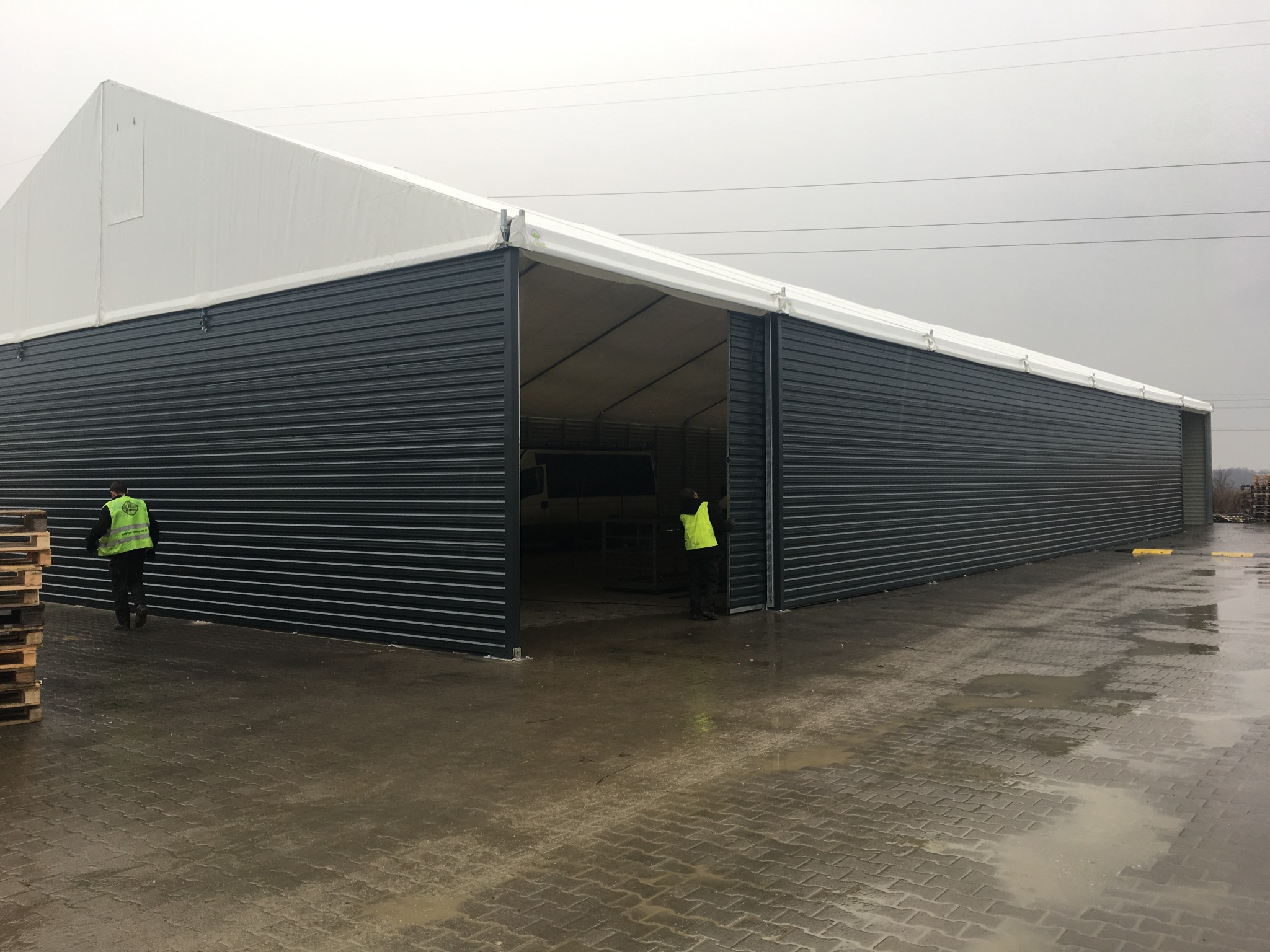 Hala z płyt warstwowych w szary odcieniu, z otwartymi wrotami, przez które widać zaparkowany samochód dostawczy. Dwóch pracowników w kamizelkach odblaskowych.