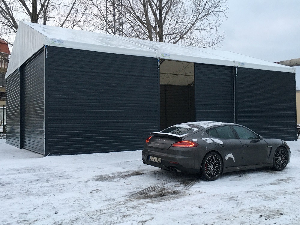 Hala z płyt warstwowych w zimowej scenerii, z częściowo otwartymi przesuwnymi drzwiami i zaparkowanym obok samochodem Porsche Panamera. Widoczne ślady opon na śniegu.