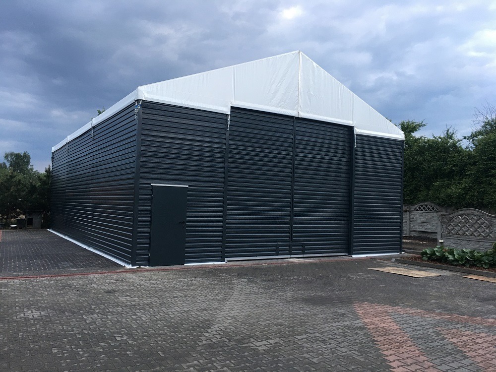 Hala z ciemnej blachy trapezowej z białym dachem namiotowym, widoczna boczna ściana z drzwiami, na pierwszym planie kostka brukowa, w tle zieleń.