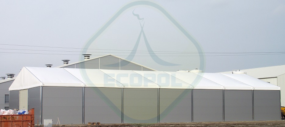 Hala namiotowa z szarymi ścianami i białym dachem, widok z zewnątrz. Na dachu widoczne zbiorniki. W tle inne budynki z kominami.