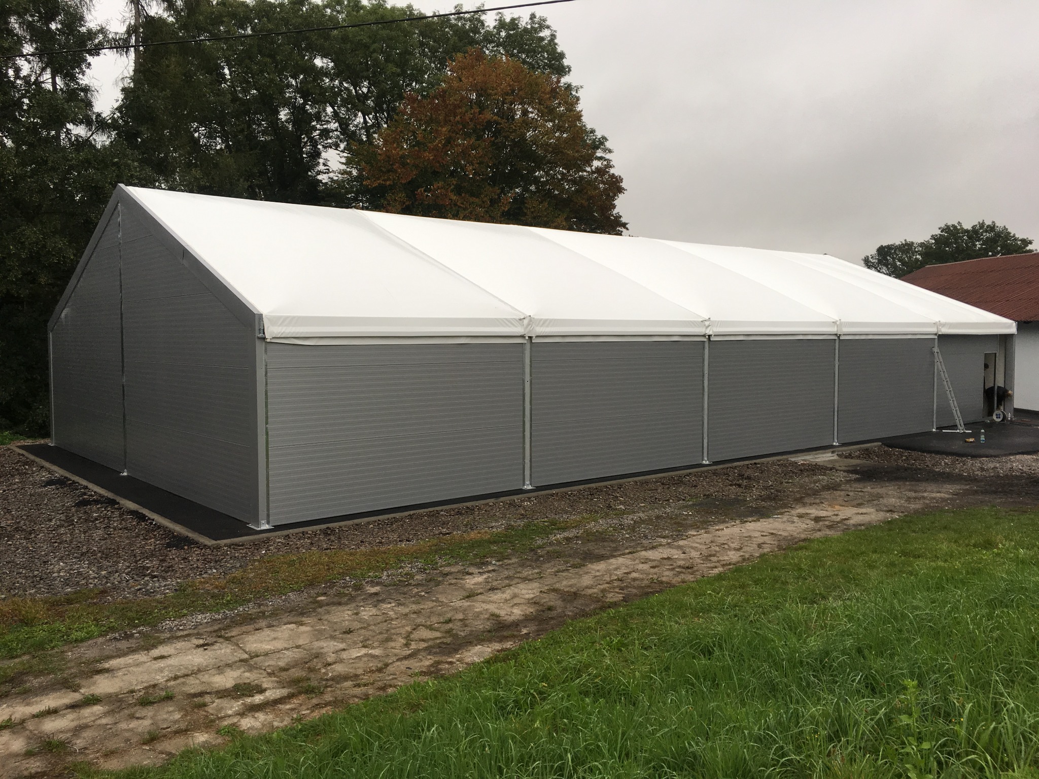 Gotowa hala z płyt warstwowych, szara elewacja, biały dach, widok z zewnątrz, teren zielony, pochmurne niebo. Widoczna drabina oparta o ścianę.