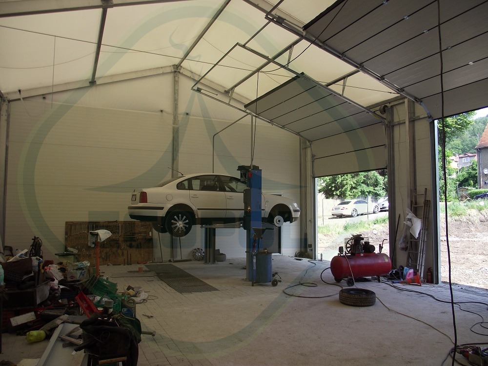 Wnętrze hali z podnośnikiem samochodowym i białym autem. Widoczne płyty warstwowe na ścianach i konstrukcja stalowa dachu. Otwarta brama wjazdowa.