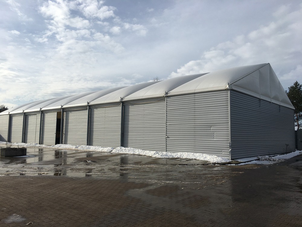 Szara hala stalowa z białym dachem na tle pochmurnego nieba. Widoczne fragmenty śniegu i kałuże na brukowanym placu przed halą. Zamknięte, metalowe bramy wjazdowe.