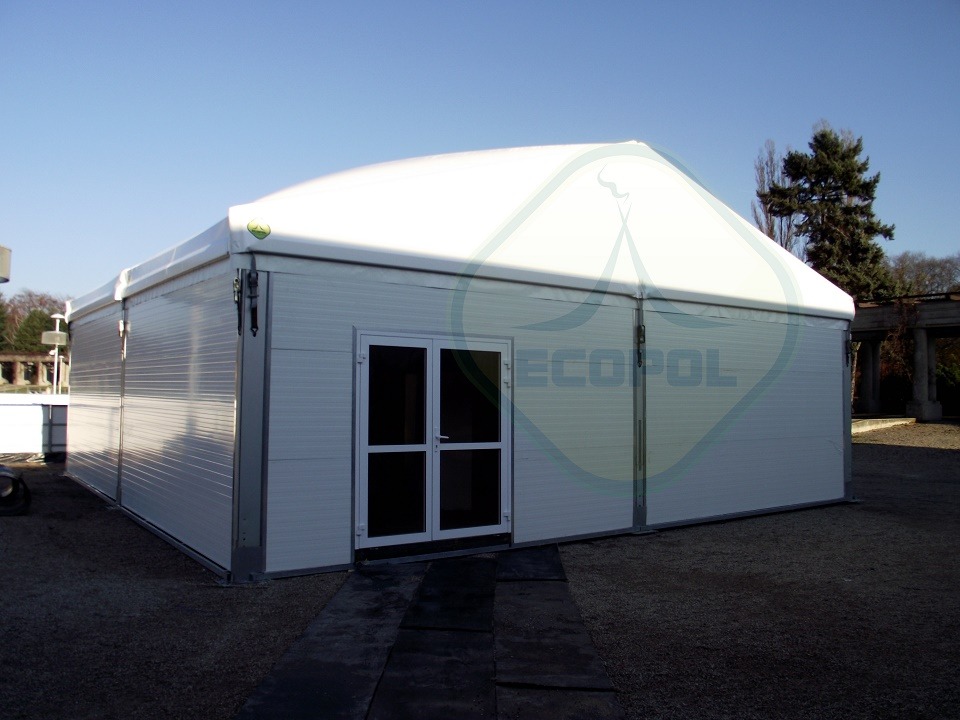 Biała hala namiotowa z dwuskrzydłowymi drzwiami wejściowymi, widoczna z zewnątrz na tle błękitnego nieba i drzew. Konstrukcja z paneli z delikatnym żłobieniem.