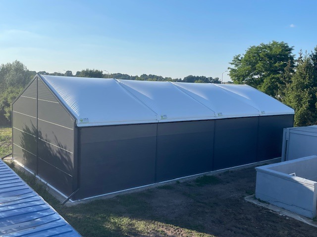 Zmontowana hala z szarej płyty warstwowej i białym dachem membranowym, widoczna z zewnątrz w słoneczny dzień. Widok z boku, z fragmentem niebieskiego dachu w lewym dolnym rogu.