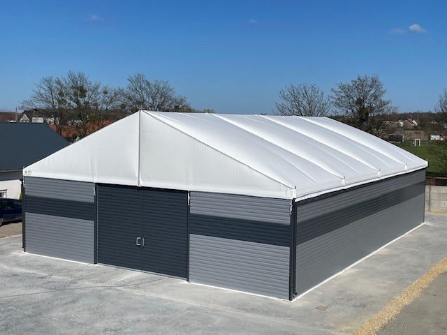 Gotowa hala z płyt warstwowych i białym dachem, widoczna z zewnątrz w słoneczny dzień. Szara brama przesuwna. Widoczne drzewa w tle.