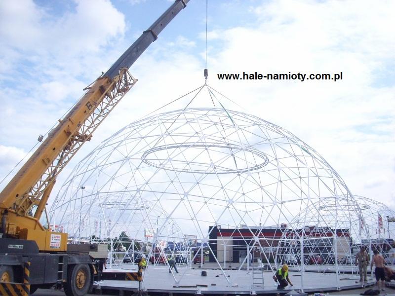 Żuraw budowlany podnosi element konstrukcyjny kopuły geodezyjnej na placu budowy, widoczni pracownicy w kamizelkach odblaskowych, w tle budynek z czerwonym dachem i adres strony internetowej...