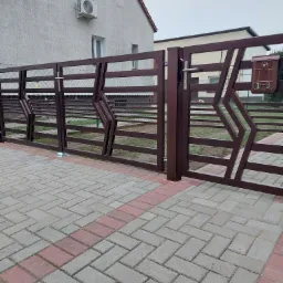 Nowoczesna, brązowa brama wjazdowa z geometrycznym wzorem i skrzynką na listy z numerem domu, widok z zewnątrz na kostkę brukową.