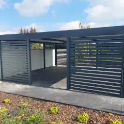 Nowoczesna, szara wiata garażowa z poziomymi panelami, wykończona kostką brukową i rabatą z roślinami ozdobnymi, na tle błękitnego nieba z chmurami.