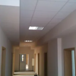 Długi korytarz w trakcie wykańczania, z podwieszanym sufitem kasetonowym z wbudowanym oświetleniem, świeżo pomalowanymi ścianami i widoczną surową podłogą przed położeniem wykończenia.