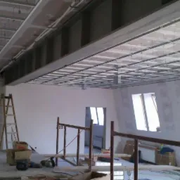 Sufit podwieszany z metalową konstrukcją w trakcie montażu, widoczne profile i elementy mocujące, surowe ściany w tle, okna w skośnych wnękach.