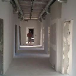 Długi korytarz w trakcie remontu z widocznymi instalacjami pod sufitem, częściowo wykończonymi ścianami i robotnikiem w oddali.