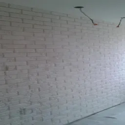 Ściana wykończona białą cegłą dekoracyjną w nowym wnętrzu. Widoczne przewody wystające z sufitu i fragment okna. Minimalistyczny styl wykończenia.