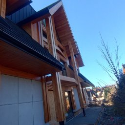 Artbudarturdudzik - Nowoczesny dom w stylu góralskim w trakcie budowy. Widoczne drewniane elementy konstrukcyjne i surowe ściany. Ujęcie z dołu, błękitne niebo w tle.