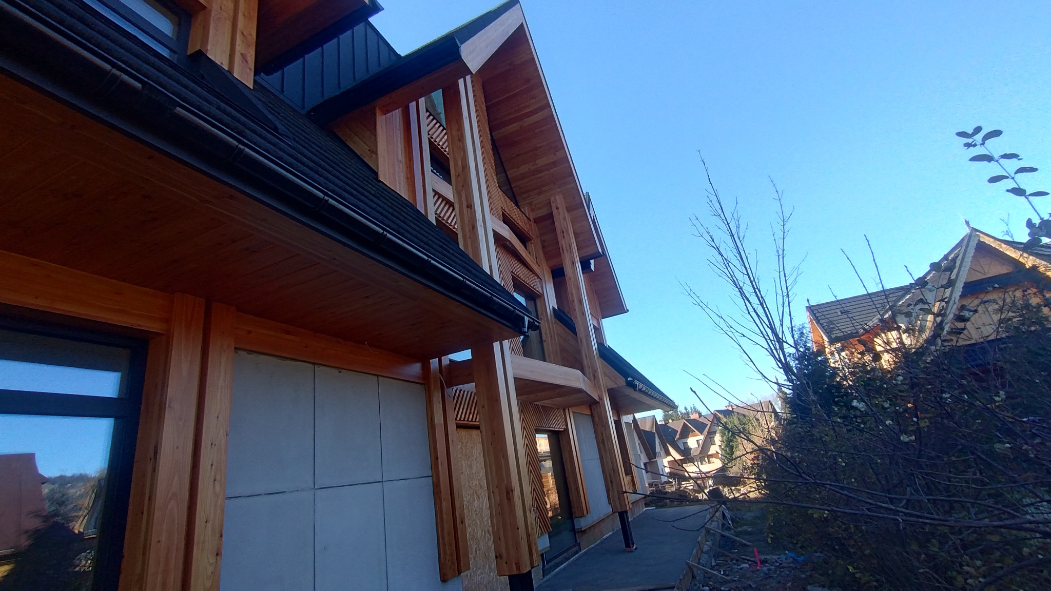 Nowoczesny dom w stylu góralskim w trakcie budowy. Widoczne drewniane elementy konstrukcyjne i surowe ściany. Ujęcie z dołu, błękitne niebo w tle.