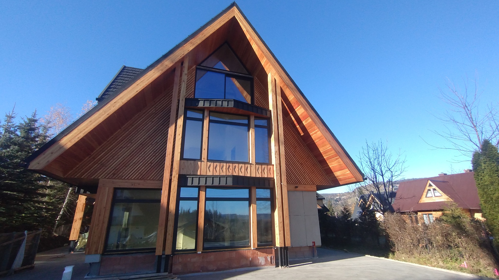 Nowoczesny dom z drewnianą elewacją i dużymi oknami, widok z dołu na tle błękitnego nieba. Charakterystyczny trójkątny szczyt i geometryczny układ okien.