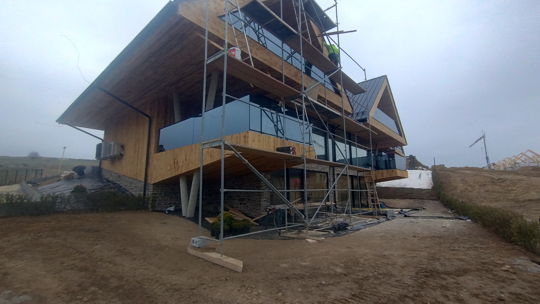 Nowoczesny, drewniany dom w trakcie budowy, z rusztowaniami, widok z dołu, pochmurne niebo. Elementy wykończeniowe i krajobraz w tle.