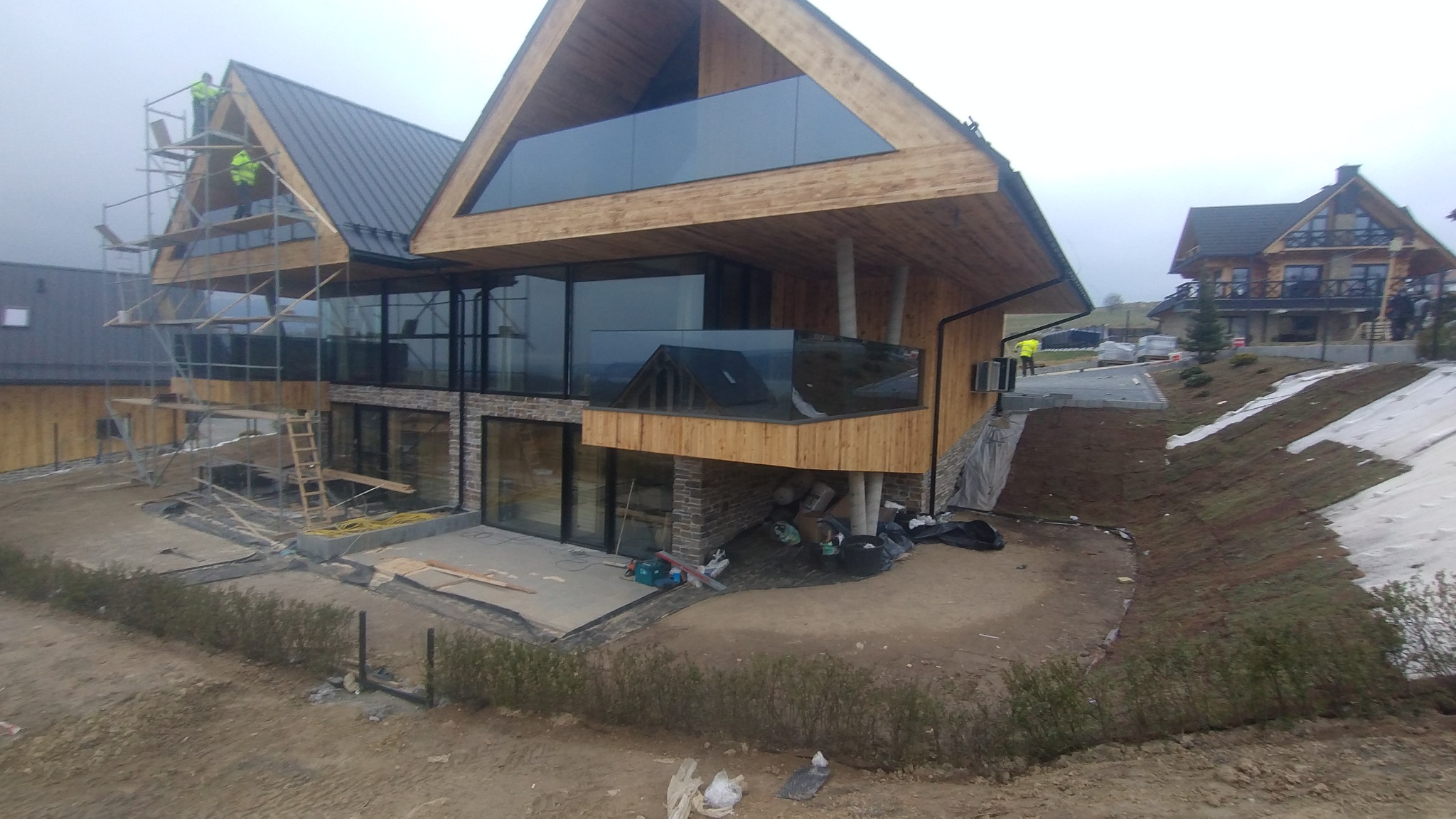 Nowoczesny dom w trakcie budowy, z drewnianą elewacją, dużymi przeszkleniami i metalowym dachem. Widoczne rusztowania i prace wykończeniowe.