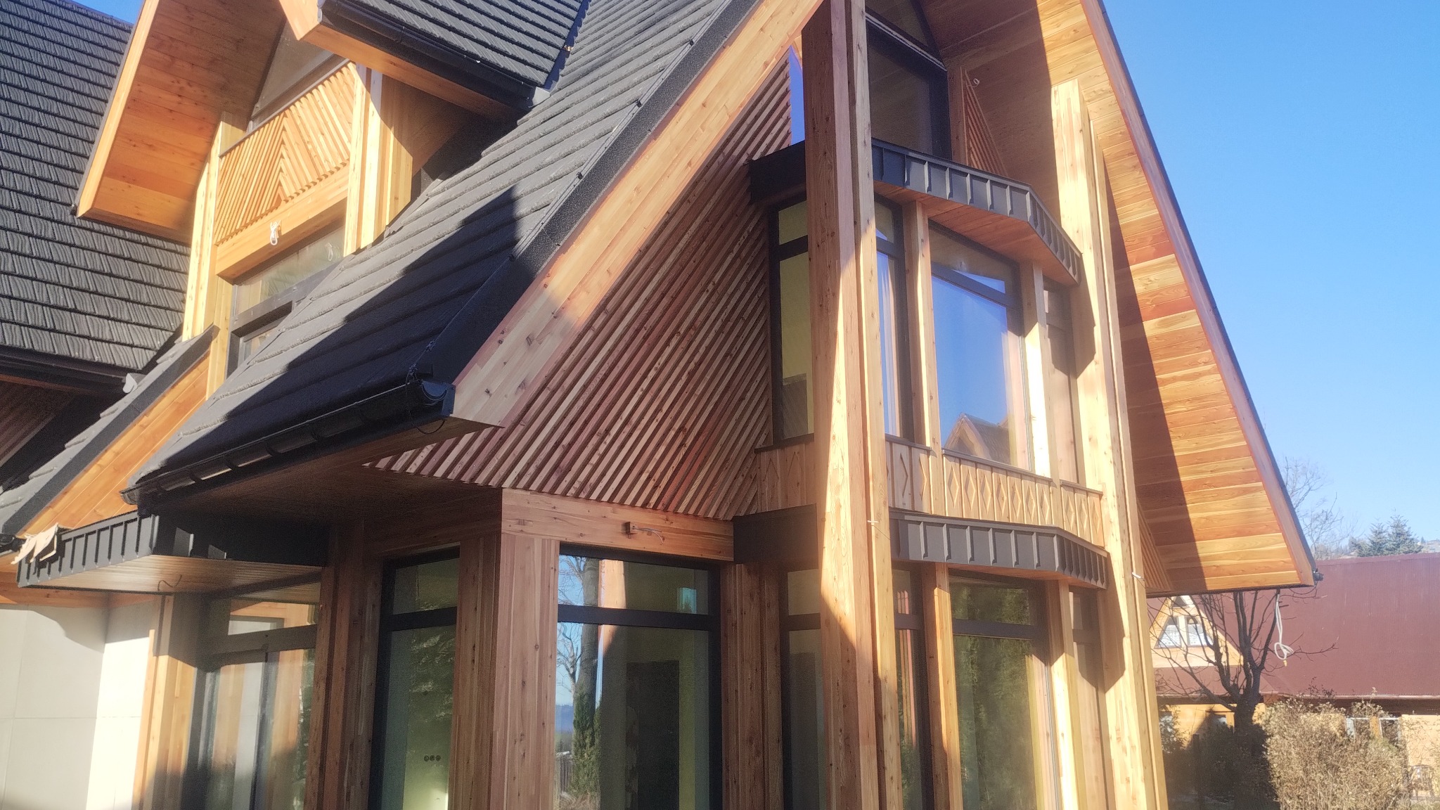 Nowoczesny dom z drewnianą elewacją w stylu góralskim, z dużymi oknami i ciemnym dachem. Charakterystyczne skośne deski i jasne drewno tworzą unikalny wygląd.