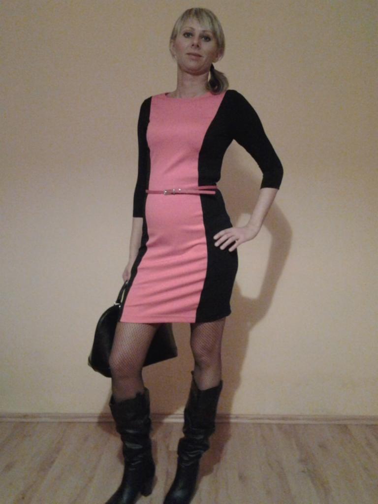 Kobieta prezentuje sukienkę w kolorze różowo-czarnym z cienkim paskiem, czarne kozaki i torebkę, na tle ściany w ciepłym odcieniu.