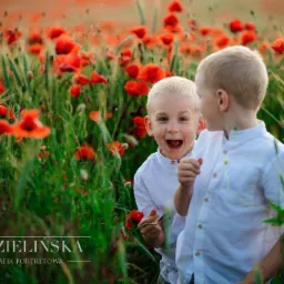 Dwóch blond chłopców w białych koszulach stoi na łące z czerwonymi makami, jeden z otwartą buzią trzyma mak
