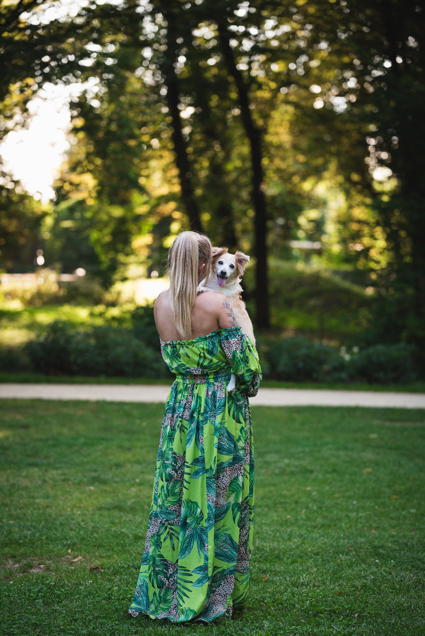Kobieta w zielonej sukience z motywem liści trzyma psa na rękach, stojąc na trawniku w parku. Pies wystawia język.
