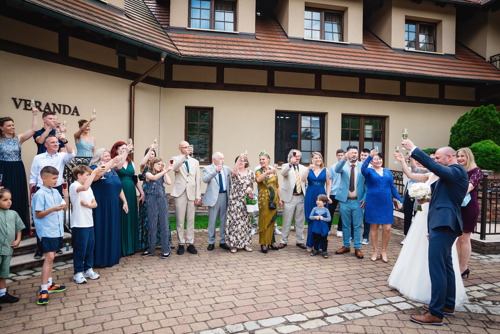 Grupa gości weselnych wznosi toast szampanem przed budynkiem Veranda w Zielonej Górze; panna młoda w białej sukni i pan młody w granatowym garniturze w centrum kadru.