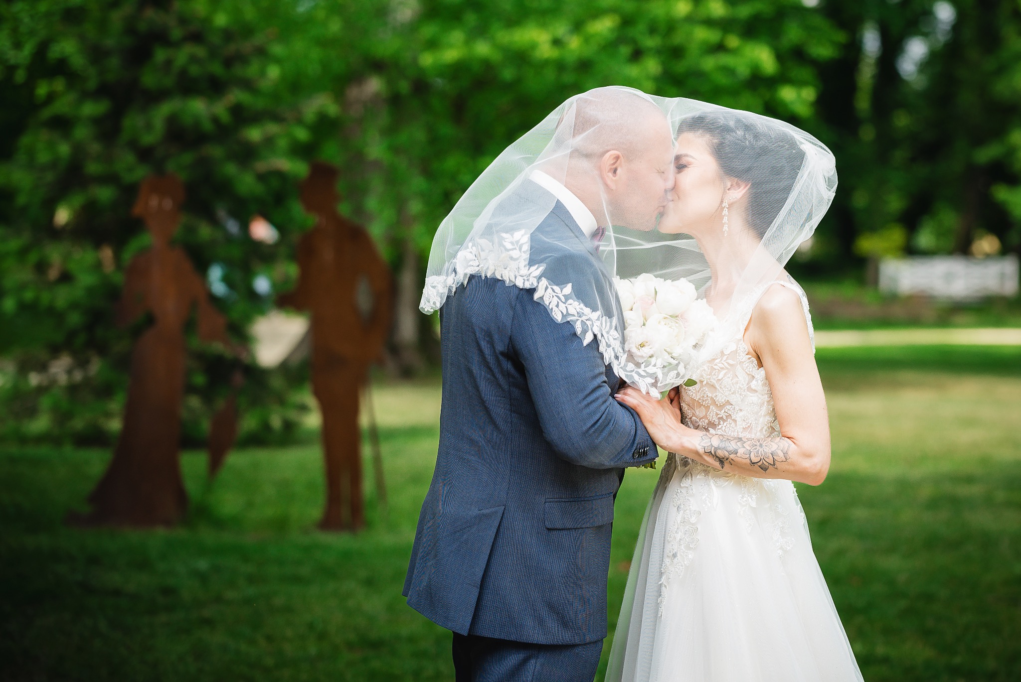 Pocałunek pary młodej pod welonem w plenerze, w tle metalowe rzeźby postaci na tle zieleni.