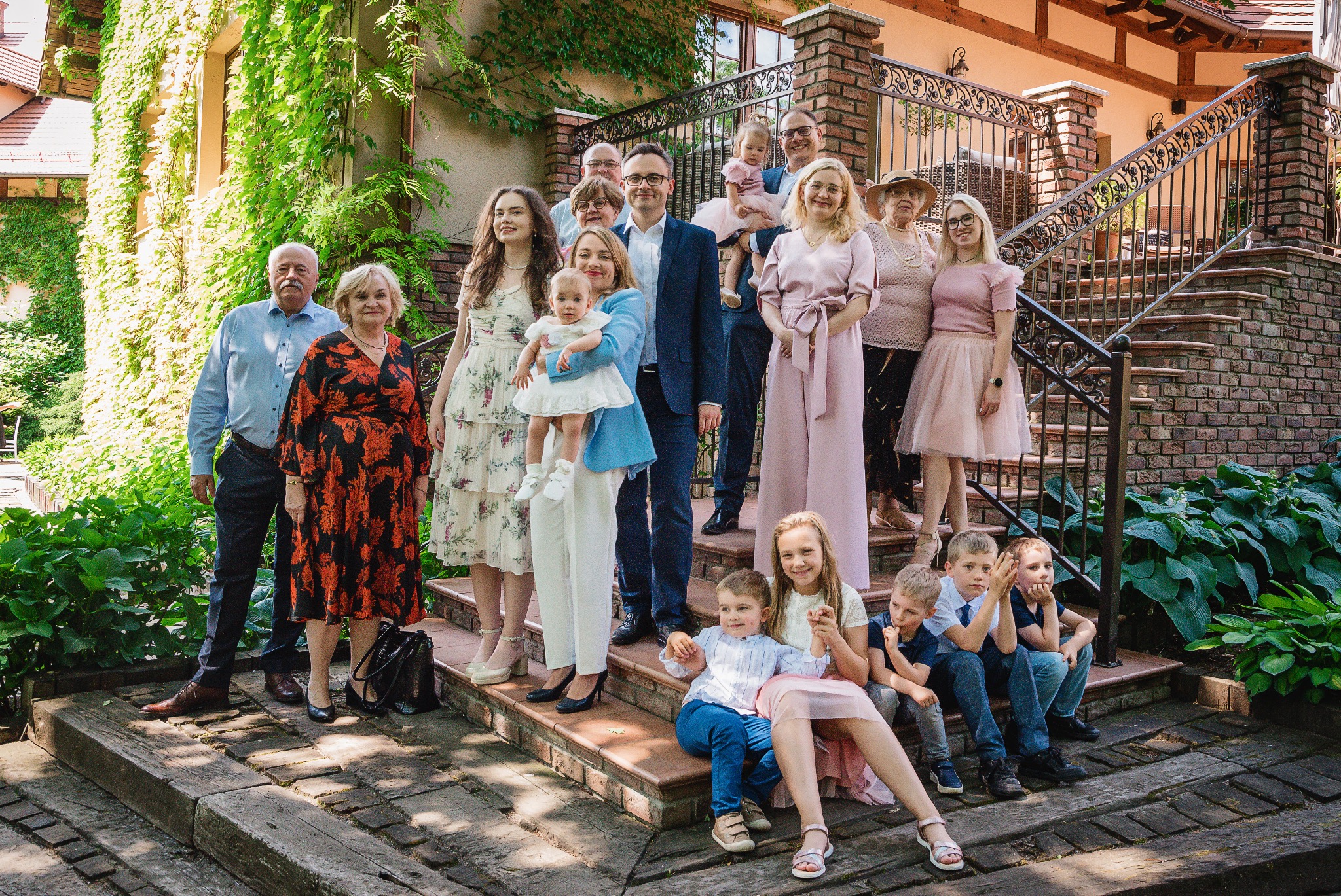 Wielopokoleniowa rodzina pozuje do zdjęcia na schodach z cegły, otoczonych zielenią, w słoneczny dzień. Dzieci siedzą na stopniach, dorośli stoją na wyższych poziomach.