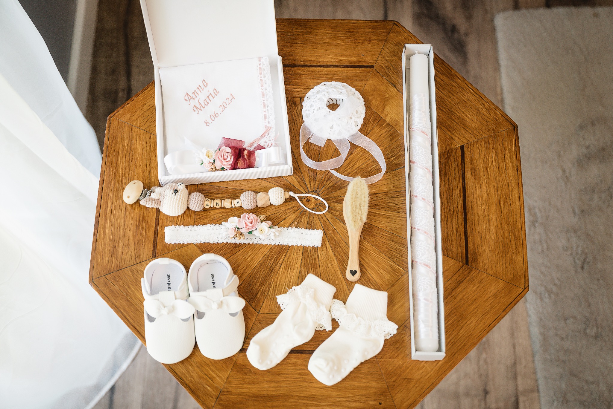 Komplet akcesoriów do chrztu na drewnianym stoliku: białe buciki, skarpetki z koronką, świeca w pudełku, szczotka z serduszkiem, smoczek z imieniem 'Ania', opaska z kwiatami i pudełko z napisem...