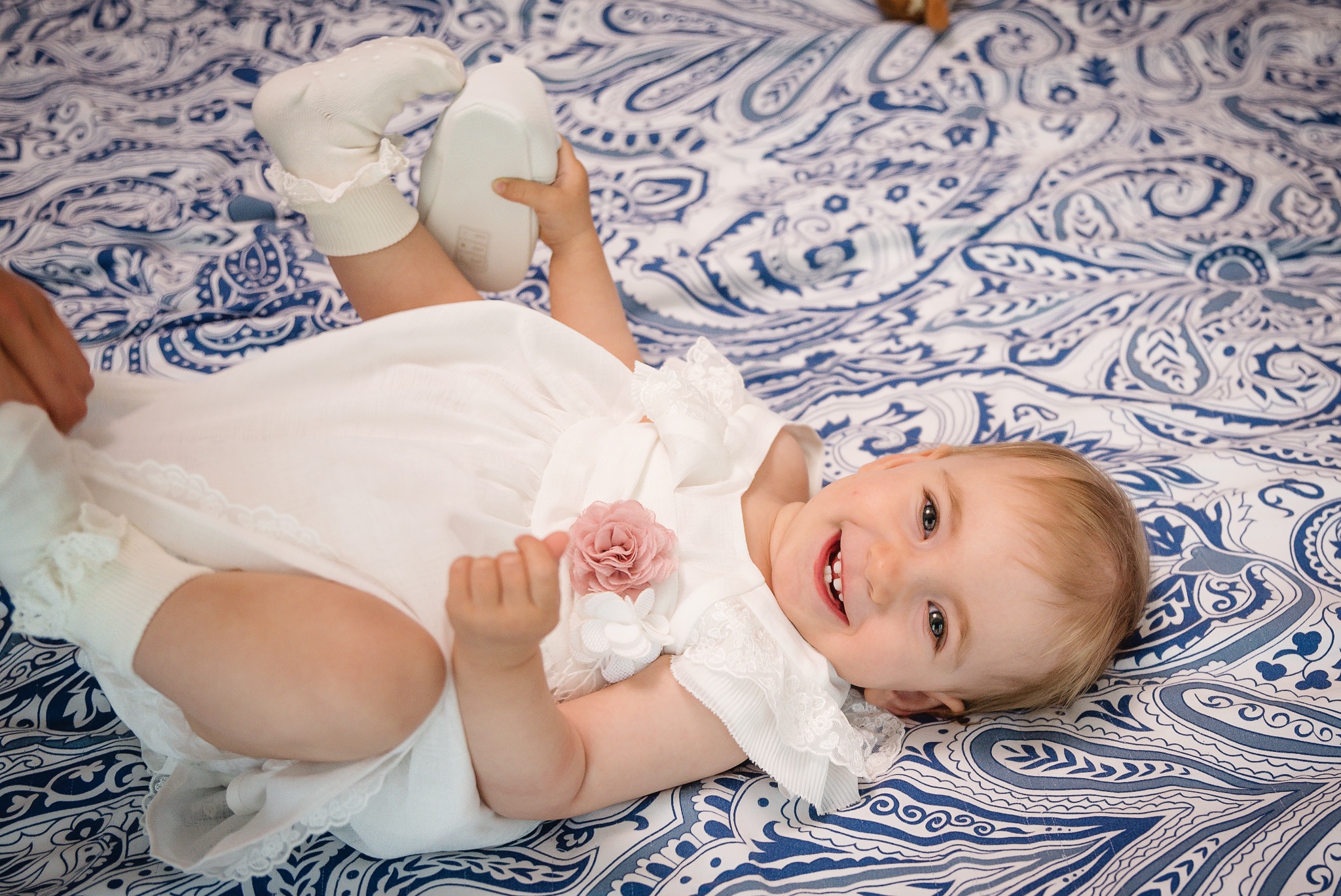 Uśmiechnięta dziewczynka w białej sukience z różową ozdobą, leżąca na plecach na niebiesko-białym wzorzystym kocyku, trzymająca w ręce biały but.